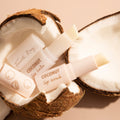 Coco Kisses ColourPop Lip Care Set showing Fourth Ray Beauty coconut lip balm and lip scrub sticks, uncapped and angled, cradled in cracked coconut halves on a soft beige surface, highlighting a tropical, hydrating lip care duo.