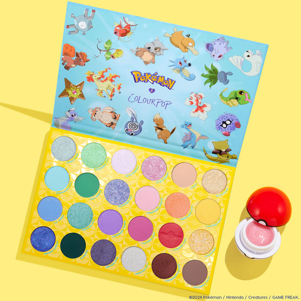 Gotta Catch ’Em All!™ ColourPop eyeshadow palette opened to show 24 vibrant matte and shimmer pans with Pokémon art on the inner lid, alongside a Poké Ball–shaped pot of pink gloss, set on a bright yellow backdrop.