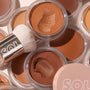 ColourPop SOL Face & Body Bronzing Balm in Fair, creamy bronzer with a fresh swipe in a clear jar, shown with a SOL white kabuki brush among open pots in tan to deep bronze shades in a studio flat lay.
