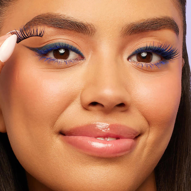 Bright cobalt lashes coated in Blue Ya Mind ColourPop BFF Mascara fan out on a model's eyes as she lifts a false lash near her lid; bold blue liner and glossy nude lips finish the studio close-up beauty look.