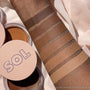 ColourPop SOL Face & Body Bronzing Balm in Medium, a warm tan cream, swatched on a forearm between Medium Dark and Light, with labeled stripes of all shades (Deep Dark to Light) beside open SOL Body compacts on soft fabric.