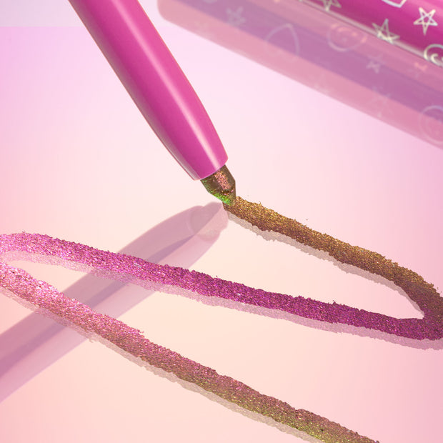 Pixie chrome crème gel liner gliding across a glossy pink surface, leaving duochrome strokes shifting from fuchsia to gold‑green, with the pink pencil barrel and star‑printed cap nearby; ColourPop Chrome Crème Gel Liner.