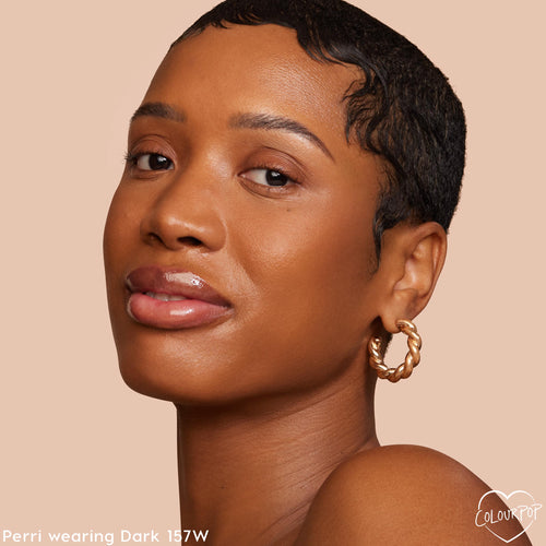 ColourPop Hyaluronic Hydrating Foundation in Dark 157 W evening the model’s complexion with a dewy, skin-like finish; close-up headshot shows smooth coverage, glossy lips, and a gold twisted hoop earring against a soft beige studio backdrop.