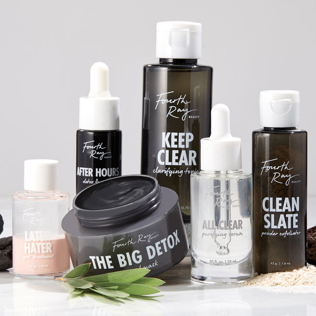 The Big Detox Mud Mask jar, lid off revealing charcoal-gray clay, sits centered among Fourth Ray Beauty skincare bottles on a white counter with green leaves; a ColourPop Face Mask for deep pore detox and clarifying.