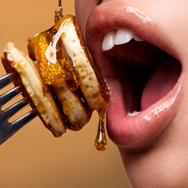 Glossy, nude lips coated in Colourpop Fresh Kiss Glossy Balm in Buttery Pancakes shade hover near syrup-drenched mini pancakes on a fork, highlighting the high-shine Lip Gloss & Lip Oil finish against a deep red background.