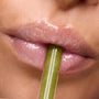 Close-up glossy lips wearing Colourpop Fresh Kiss Glossy Balm Vanilla Matcha shade, holding a shimmery green lip applicator between them, promoting hydrating Lip Gloss & Lip Oil finish 
