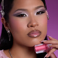 Model wears ColourPop Your New Obsession ultra glossy lip from the Heart to Get collection, showing high-shine nude lips while holding the pink gloss tube near her face against a bold magenta background.