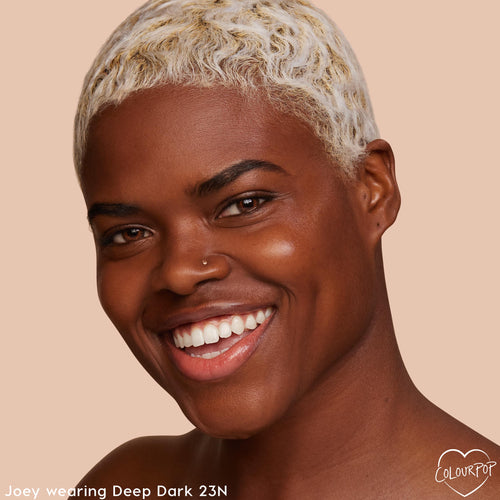 Model wearing Deep Dark 23 N smiles, showing smooth, radiant skin from the ColourPop Pretty Fresh Tinted Moisturizer; close-up face with short bleached waves on a warm beige studio background.