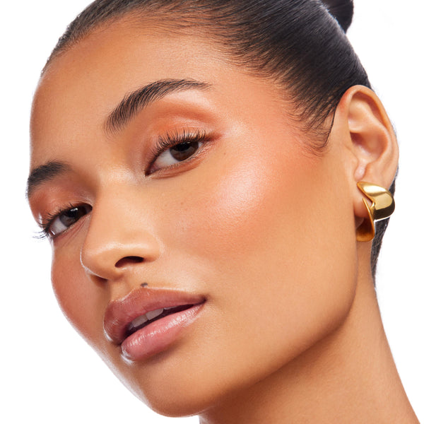 Ignite Lite Stix cream highlighter from ColourPop Lite Stix sweeping a peachy-gold glow across a model’s cheekbones and brow bone, catching light in a close-up with glossy lips and a gold earring against a clean white background.