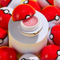 You're a Catch ColourPop Lip Mask - pink, glossy balm - sits open in a Poké Ball–style red-and-white container on a pastel pedestal, surrounded by matching spheres under bright studio light, highlighting the playful packaging and lip care product.