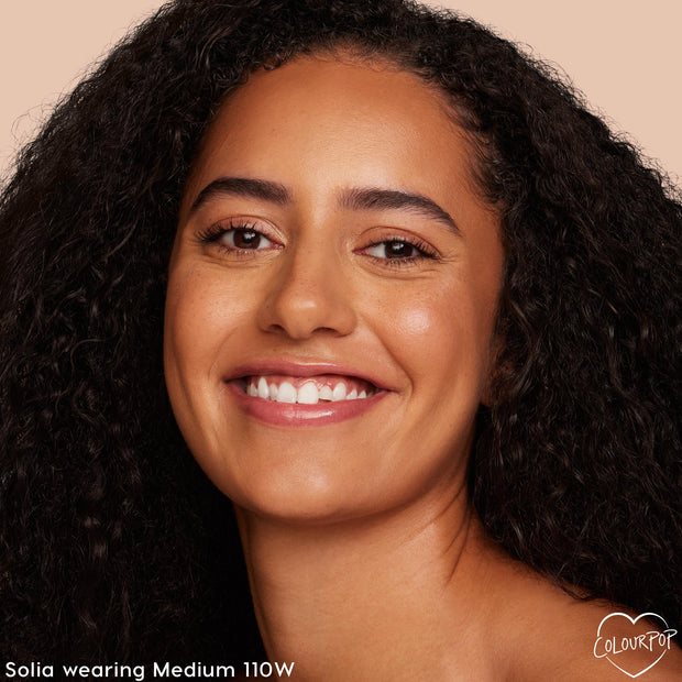 Model smiling while wearing ColourPop Hyaluronic Hydrating Foundation in Medium 110 W, showcasing smooth, dewy coverage with a warm medium undertone in a studio close-up against a neutral beige background.