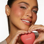 ColourPop Heart Powder Blush Set Totally Lovestruck compact held between a model’s hands under her smiling face, showcasing soft rosy cheeks; coral heart-shaped case with ColourPop logo in a clean studio close-up.