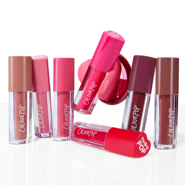 ColourPop Lip Stain Pop Stars tubes standing, leaning, and one lying to show nude, pink, and berry shades, paired with a hot‑pink Hydra Pop PopSocket phone grip, arranged on a glossy white studio surface.
