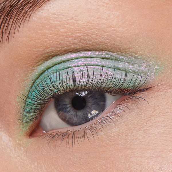 Eyelid wears Solar Beam, a ColourPop Super Shock Shadow, shifting mint green to pink‑violet iridescent shimmer across the lid, defining the crease and lash line with fine sparkle in a close-up eye view.