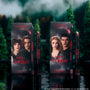 ColourPop Twilight Immortal Kiss Lippie Stix Duo stands upright on a reflective lake surface, with Eclipse-themed character boxes and matching deep nude and berry lip pencils and sticks framed by a dark, misty forest backdrop.