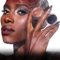 Model with dramatic smokey eye and deep berry lips holds two Twilight Coven Clash Jelly Much Duo pots, one copper shimmer and one inky black, near her face in a smoky, high-fashion beauty close-up.