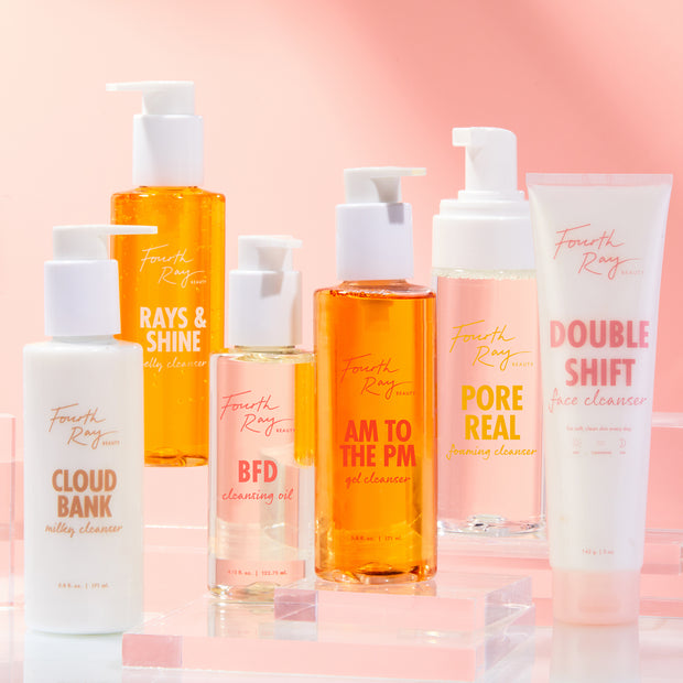 AM to the PM Gel Cleanser by ColourPop Fourth Ray Beauty stands centered, an amber gel pump bottle showcased on clear acrylic risers, surrounded by other Fourth Ray face cleansers against a pink gradient background.