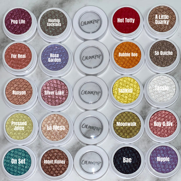 La Mesa shimmery peach‑champagne ColourPop Super Shock Shadow displayed in a white round pot, its bouncy textured metallic finish catching light, arranged in a flatlay grid with other shadow pots on a marble countertop.