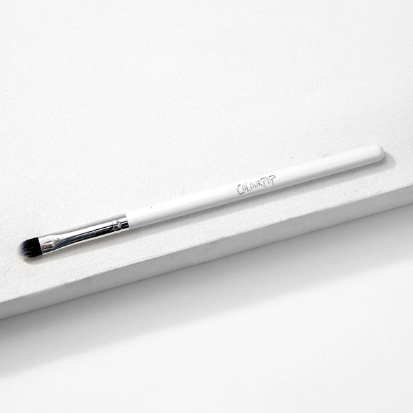 ColourPop Small Shader Brush makeup brush resting diagonally on a white textured surface, showing a sleek white handle with ColourPop logo, shiny silver ferrule, and dense flat rounded bristles for precise eyeshadow application.
