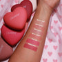 ColourPop Kiss 'N Tell pressed powder blush swatched on a forearm, with six labeled shades - Kiss N Tell, Let’s Dance, Flirt Alert, 4ever Yours, Text Me, Babycakes - beside matte heart-shaped senjyuan.onlinepacts on pink heart-print fabric.