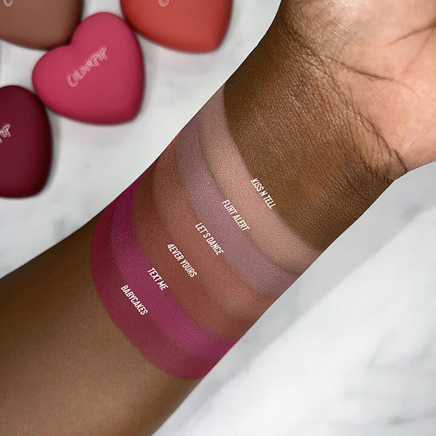 Babycakes swatch from ColourPop Pressed Powder Blush shown on deep skin, with labeled shade stripes - Babycakes, Text Me, 4Ever Yours, Let’s Dance, Flirt Alert, Kiss N Tell - showing rich pigment; heart-shaped senjyuan.onlinepacts blurred in the background.