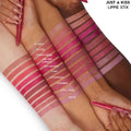 Swatched Just A Kiss ColourPop Lippie Stix shades striped on four arms, hands holding pink Lippie Stix, showing nudes to rosy pinks; labeled shades include Oh Snap, Payton, Cherry Bomb, Are You Surreal, Out of Sync, Cami, Aftershock, Songbird, Little Tings, Westie.