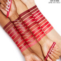 ColourPop Lippie Stix Hot in Here Vault swatches sweep across three skin tones, showing 10 reds from deep burgundy to bright coral (Infinite Best to Daytrip), as hands hold red Lippie Stix pens on a white studio background.