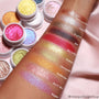 So Quiche Super Shock Shadow swatched on a forearm - shimmery olive-taupe with a green-gold shift - shown beside labeled swatches and open white pots of other ColourPop eyeshadows on a pink backdrop.