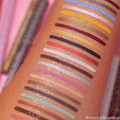 ColourPop Crème Gel Liner It’s a Vibe swatches bead water without smudging, with stacked lines of blue, yellow, gold, red, white, silver, black, and neutrals on a forearm, showing matte and metallic finishes against a pink vanity backdrop.