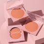ColourPop Pressed Powder Blush Shortbread compact displays a warm peach matte pan in a pale pink square case, with one boxed and two open compacts arranged on a soft pink pedestal under studio lighting.