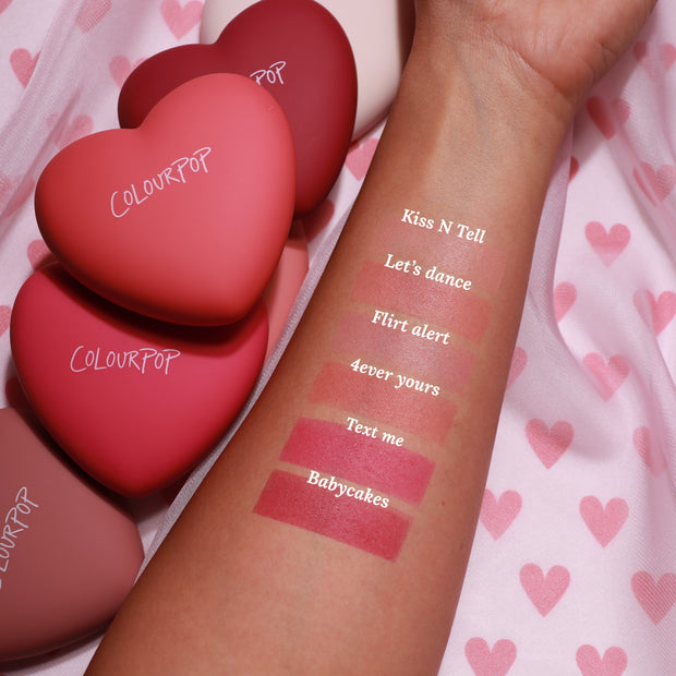 Swatched forearm displays “Text Me” ColourPop Pressed Powder Blush, a rosy pink matte stripe, alongside shades Babycakes, 4ever Yours, Flirt Alert, Let’s Dance, and Kiss N Tell; heart-shaped senjyuan.onlinepacts on pink heart-pattern fabric.