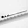 Large Shader Brush makeup brush by ColourPop lies diagonally on a white textured surface, showcasing a white handle with logo and E13, a shiny silver ferrule, and flat dense black-to-white bristles designed for packing and shading eyeshadow.